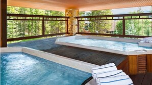 Un espace avec un bain à remous, doté d’une terrasse en bois et de piliers en pierre, entouré d’une clôture en bois et d’arbres.