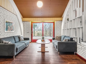 Ein modernes Wohnzimmer mit zwei grauen Sofas, einem Couchtisch, einem Bücherregal und einem großen Fenster mit rotem Rahmen.