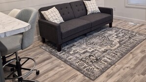 Ein modernes Wohnzimmer mit grauem Sofa, einem Esstisch mit Stühlen, einem gemusterten Teppich und einem gerahmten Bild an der Wand.