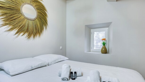 Camera d'albergo con un letto grande, lenzuola bianche, uno specchio rotondo, una piccola finestra con un vaso di fiori e articoli da toilette sul letto.