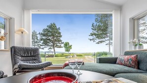 Un salon donnant sur un espace verdoyant et des arbres, avec un fauteuil en cuir, un canapé, une table sur laquelle se trouvent un bol rouge et un verre à vin, et un journal.