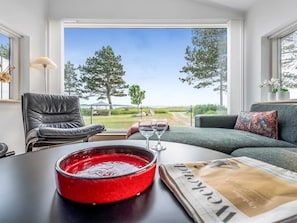 Ein Wohnzimmer mit Blick auf eine grüne Fläche und Bäume, mit einem Ledersessel, einem Sofa, einem Tisch mit roter Schüssel und Weinglas sowie einer Zeitung.