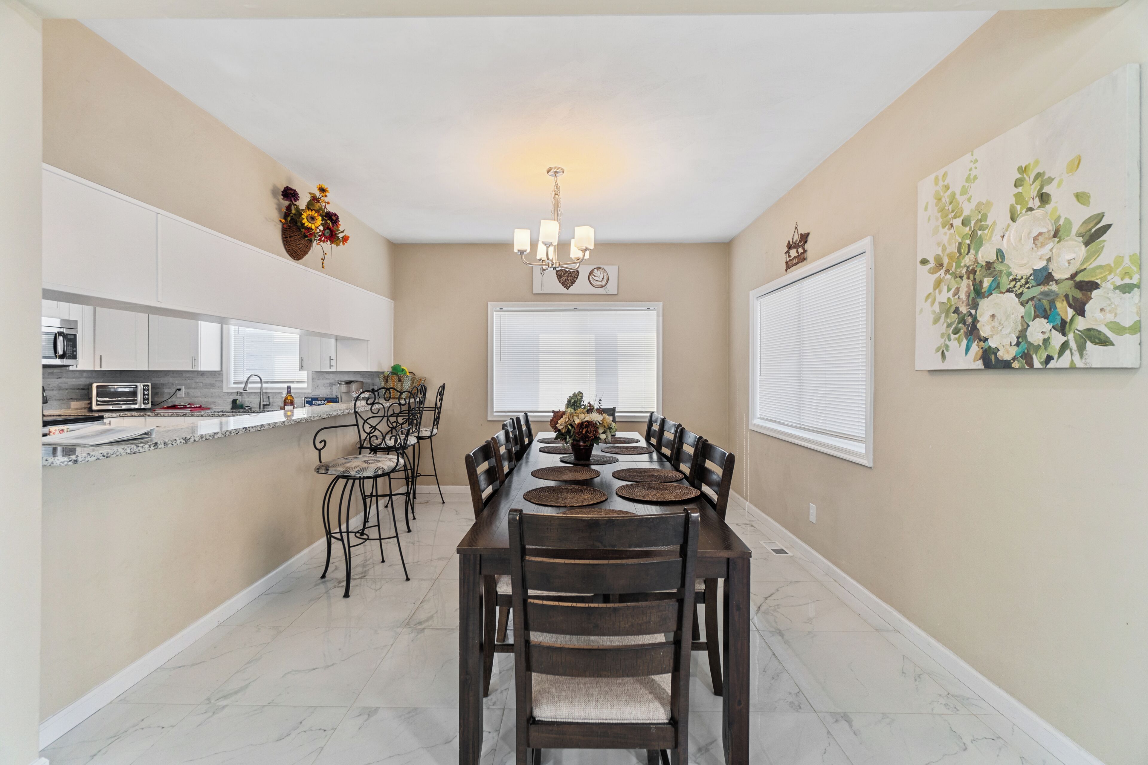 A dining area with a long table, chairs, a chandelier, and a kitchen in the background.