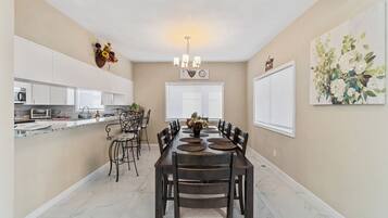 A dining area with a long table, chairs, a chandelier, and a kitchen in the background.