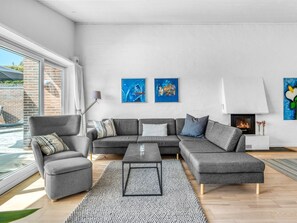 Ein modernes Wohnzimmer mit einem grauen Ecksofa, einem Couchtisch und einem Kamin. An der Wand hängen zwei abstrakte Gemälde und ein Fenster bietet Blick auf Grünflächen.