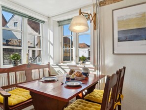 A dining area with a wooden table, yellow cushioned chairs, a hanging light fixture, and a view of buildings through large windows. - "Ariela" - 150m from the sea by Interhome (Allinge)