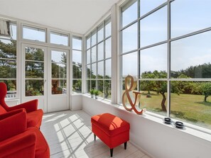 Ein Zimmer mit großen Fenstern, einem roten Sessel, einem kleinen roten Hocker und einem dekorativen Metallstück auf der Fensterbank.