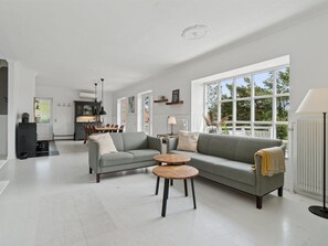 Ein geräumiges Wohnzimmer mit einem grauen Sofa, einem runden Holztisch und einem großen Fenster mit Blick auf Grünflächen.