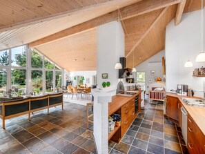 Eine geräumige Küche mit einer großen Kochinsel, ein Essbereich mit hohem Tisch und Blick ins Freie durch große Fenster.