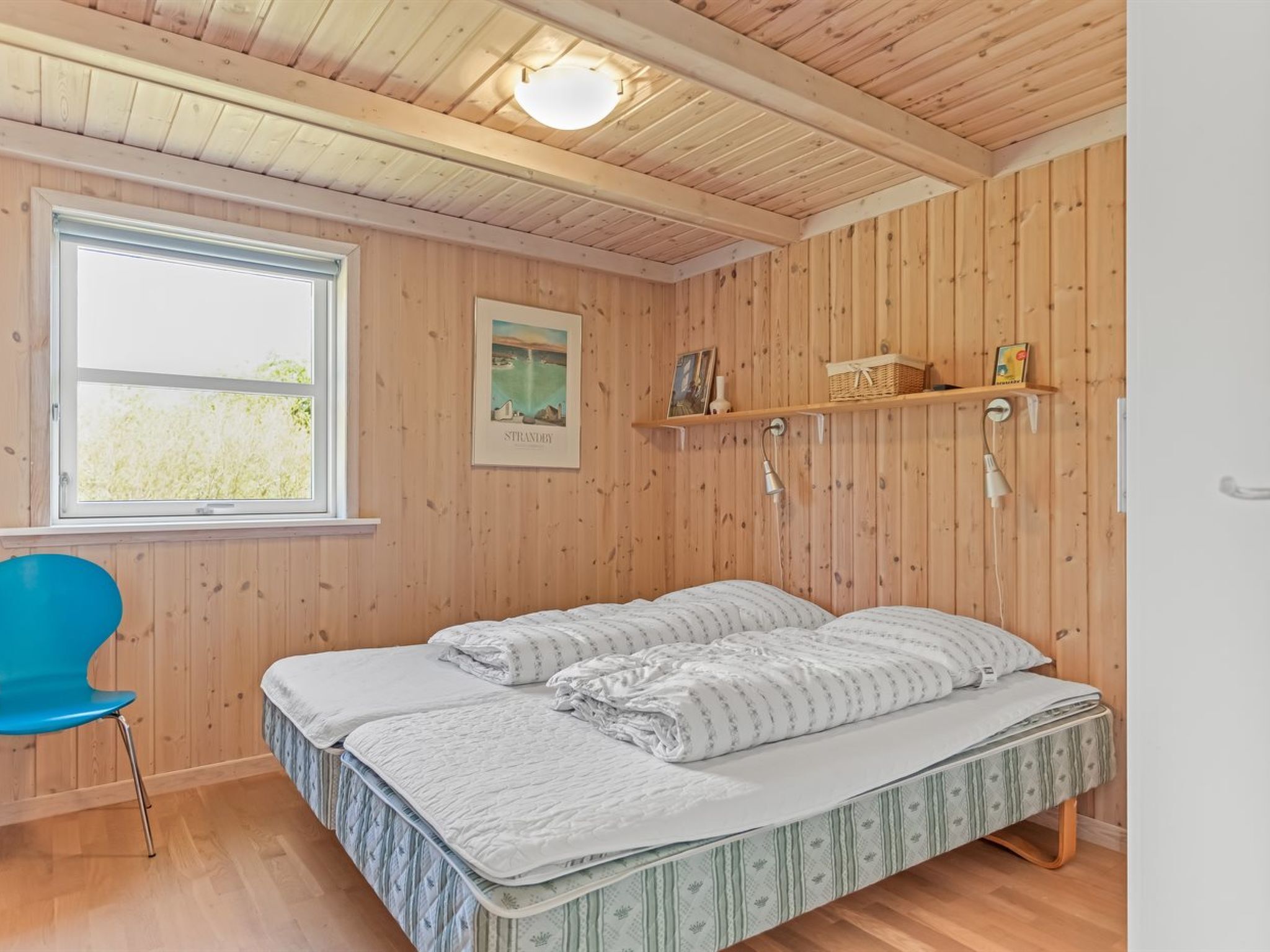 Une pièce aux murs en bois, un lit avec deux oreillers, une chaise bleue, une fenêtre et une étagère avec divers objets.
