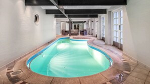 Une grande piscine intérieure à la forme incurvée, entourée d’un sol et de murs recouverts de carreaux, et dotée d’un plafond avec des poutres apparentes.