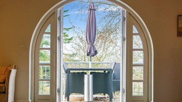 Une porte-fenêtre donnant sur un patio extérieur, avec une table et des chaises, un parasol et une vue sur les arbres et le ciel.