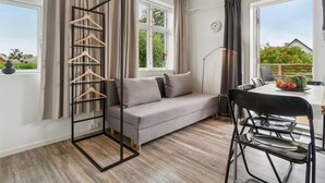 Un salon moderne avec un canapé gris, une table à manger avec des chaises, un portant à vêtements suspendu et de grandes fenêtres avec des rideaux.