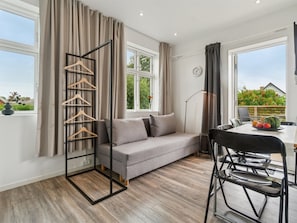 A modern living room with a grey sofa, a dining table with chairs, a hanging clothes rack, and large windows with curtains.
