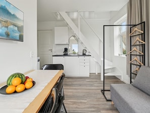 A modern kitchen with white cabinets, a black countertop, and a coffee maker. There is a dining table with a fruit bowl and a gray sofa. A staircase leads to another level.