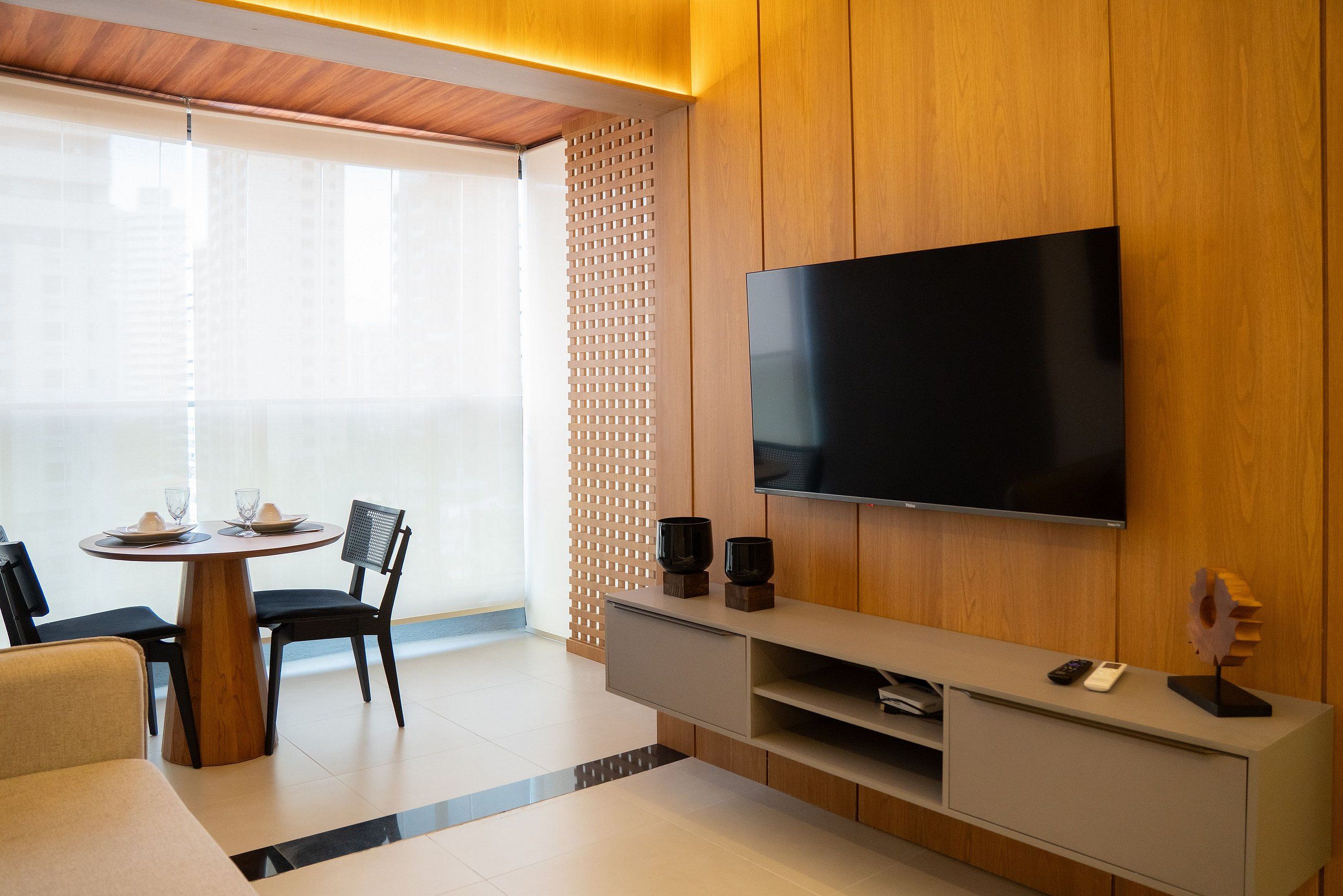 A modern living room with a flat-screen TV mounted on a wooden wall, a dining table with chairs, and a small coffee table.