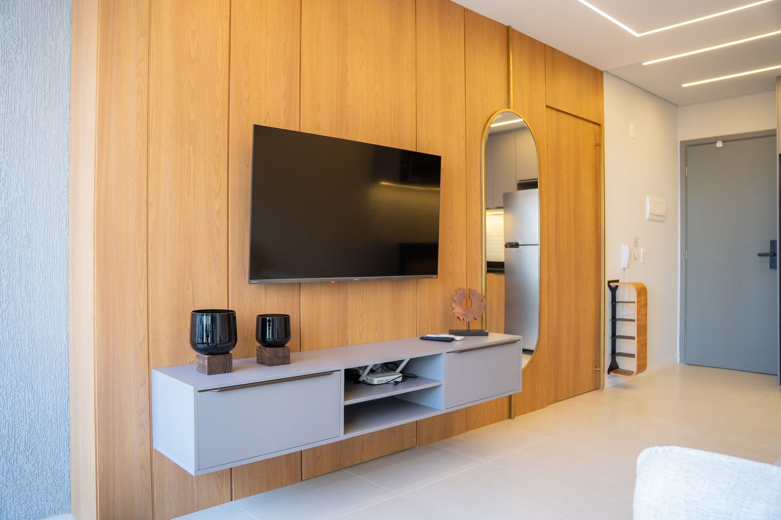 A modern living room with a flat-screen TV mounted on a wooden wall, a minimalist console table with decorative items, and a large mirror reflecting the room.