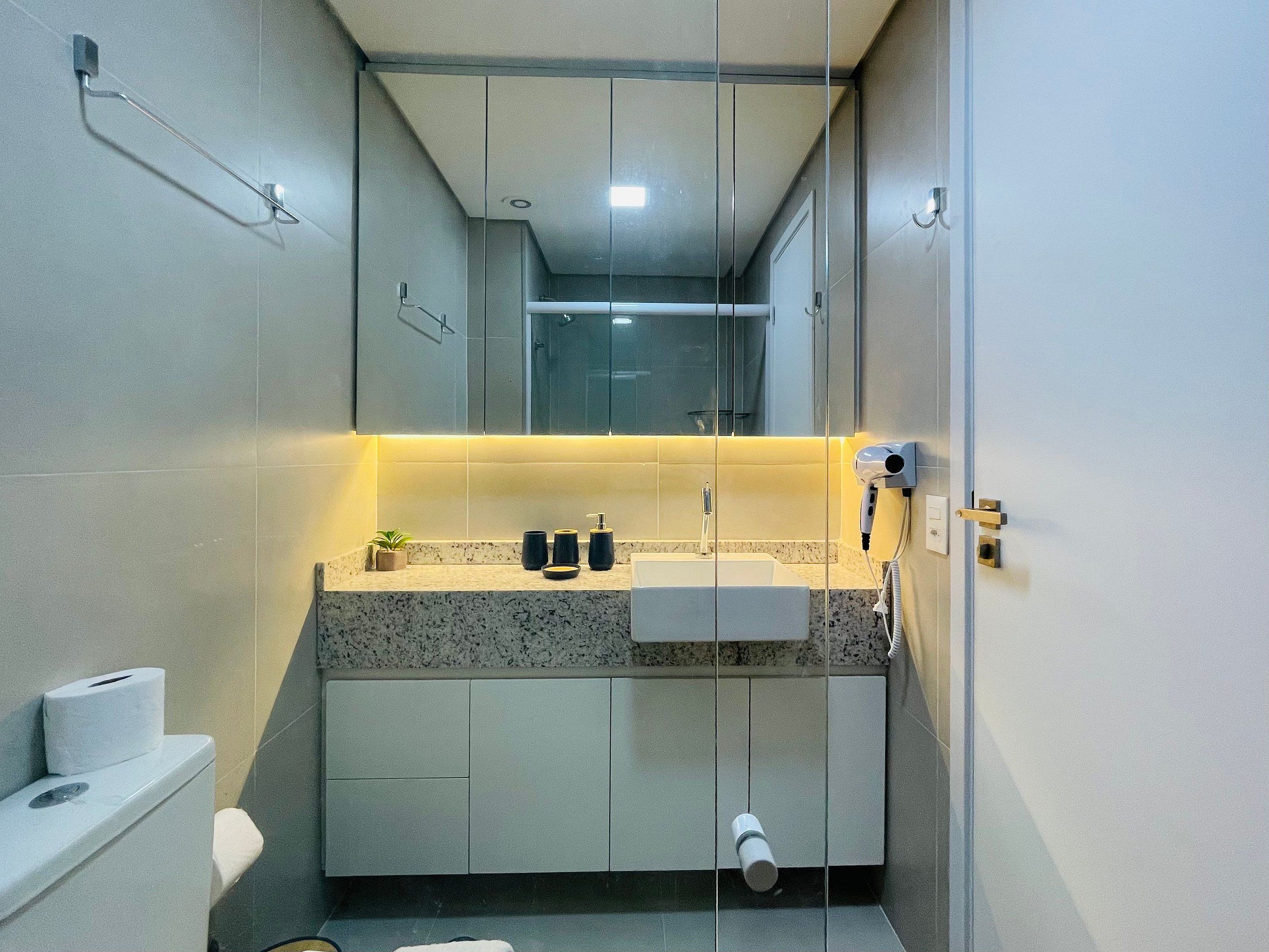 Um banheiro moderno com bancada de mármore, pia, vaso sanitário e secador de cabelo fixado na parede.