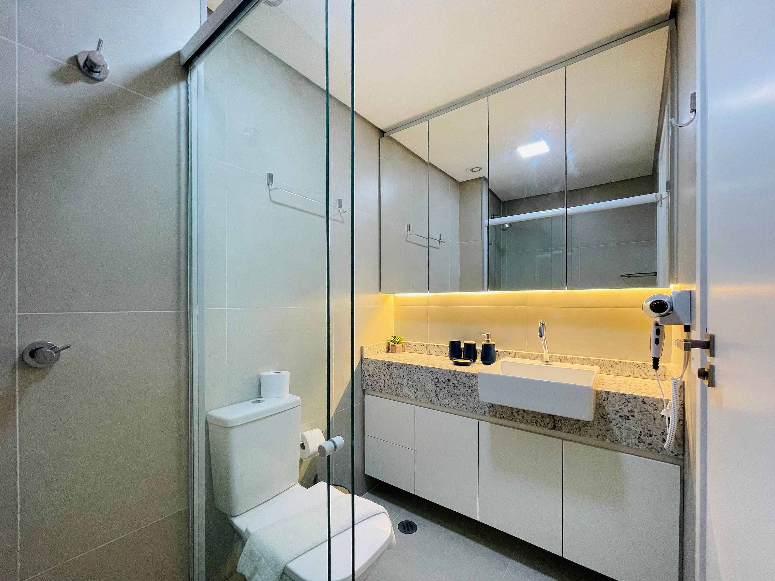 Un baño moderno con cabina de ducha de vidrio, un inodoro, un lavabo con espejo y una encimera con artículos de aseo.