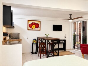 A compact living space with a kitchenette, a dining area, a TV mounted on the wall, and a balcony with a red chair. - Romantic Zone Studio · Rooftop · AC · Near Beach (Puerto Vallarta)