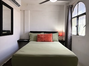 A single bed with a green bedspread and two pillows, a bedside lamp, a nightstand, a mirror, and a window with curtains. - Walkable Romantic Zone Studio • Near Beach • WiFi (Puerto Vallarta)
