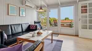 Un cuarto acogedor con un sofá de cuero negro, una mesa de centro de madera y una mesa de comedor dispuesta con platos y tazas. Hay dos cuadros enmarcados en la pared, una lámpara colgante y un ventanal corredero que da a un espacio exterior con una silla y una mesa.