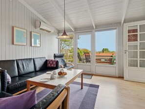 Ein gemütliches Zimmer mit einer schwarzen Ledersofa, einem Holztisch und einem Esstisch, gedeckt mit Tellern und Tassen. An der Wand befinden sich zwei gerahmte Bilder, eine hängende Pendelleuchte und eine Glastür, die zu einem Außenbereich mit einem Stuhl und einem Tisch führt.