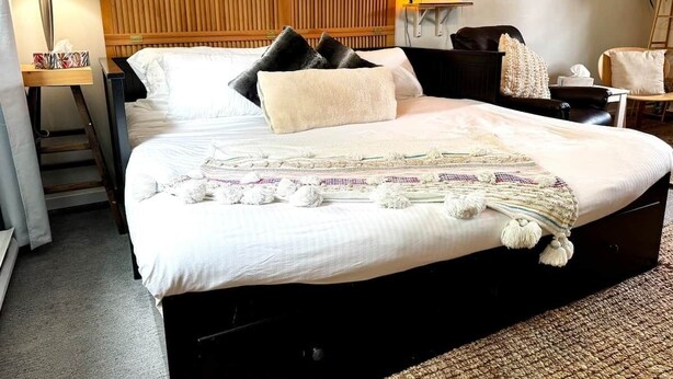 A hotel room with a large bed, a wooden headboard, a bedside table with a lamp, a chair, and a painting on the wall.