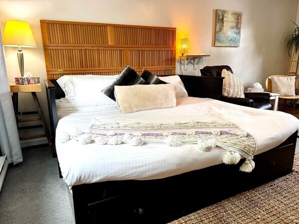 A hotel room with a large bed, a wooden headboard, a bedside table with a lamp, a chair, and a painting on the wall.