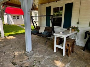 A covered patio with a swing chair, a table with a plant, and a stone floor.