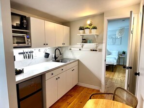 Eine kompakte Küche mit weißen Schränken, einer Mikrowelle und einem Spülbecken. Es gibt einen kleinen Essbereich mit einem Tisch und einem Stuhl.