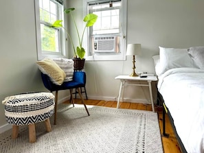 A bedroom with a bed, a chair, a small table, a plant, and a window.