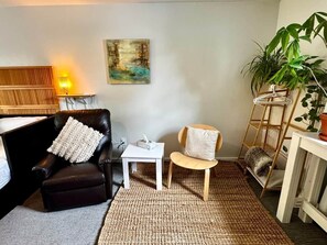 Ein gemütliches Wohnzimmer mit einer schwarzen Ledersofa, einem kleinen weißen Tisch, einem Holzstuhl, einem Gemälde an der Wand und einer Pflanze.