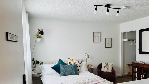 Ein Schlafzimmer mit Bett, Nachttisch, Lampe, Stuhl und einer Tür mit der Aufschrift 'STUDIO'.