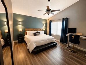 Ein Schlafzimmer mit einem Bett, Nachttischlampen, einem Deckenventilator, einem Schreibtisch mit Stuhl und einem Fenster mit Vorhängen.