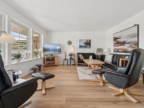 Ein modernes Wohnzimmer mit großem Fernseher, schwarzen Ledersesseln, einem Holztisch und Blick auf ein Gebäude durch große Fenster.