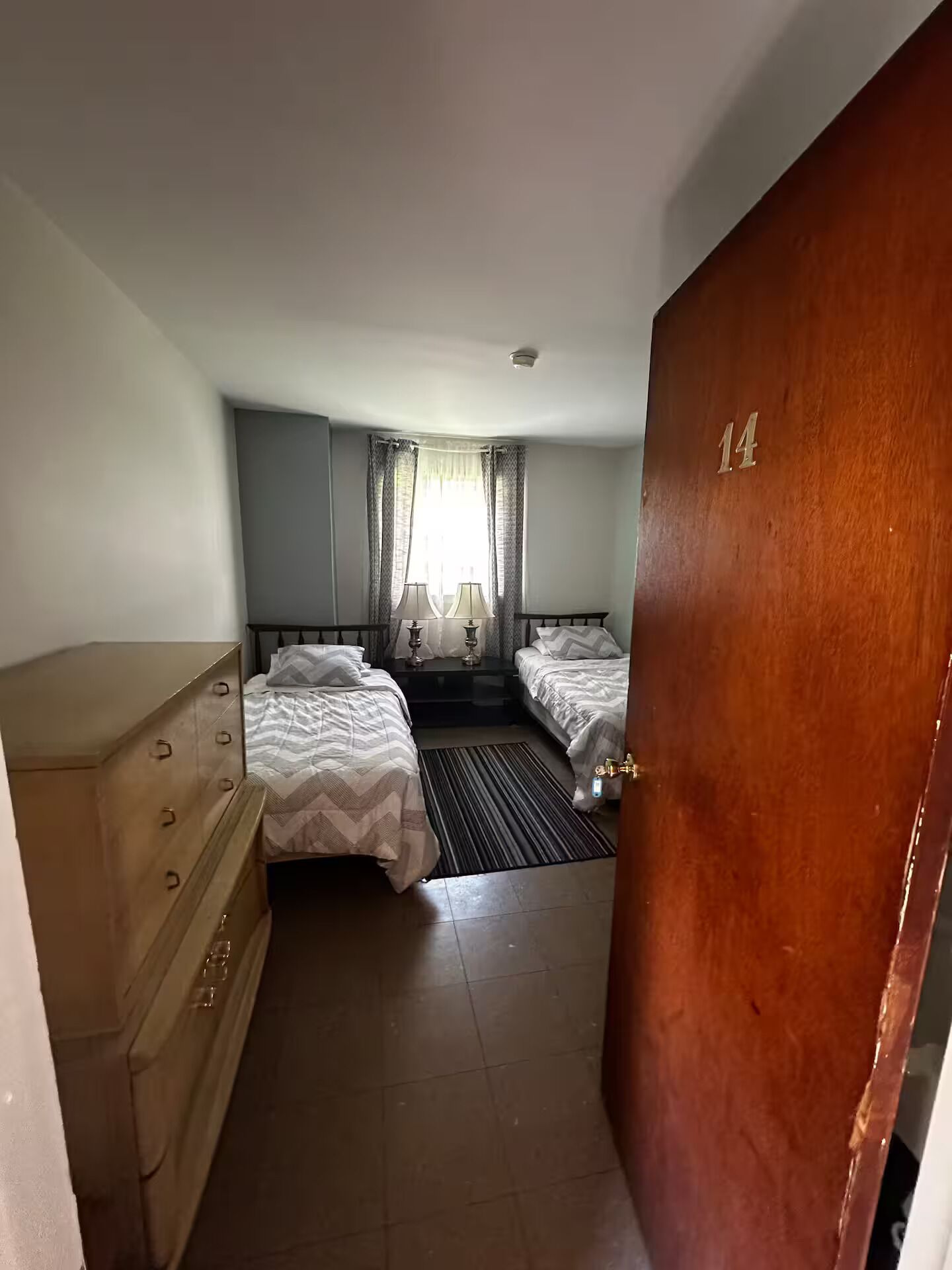 Habitación de hotel con dos camas, un aparador de madera, una ventana con cortinas y una puerta con el número 14.
