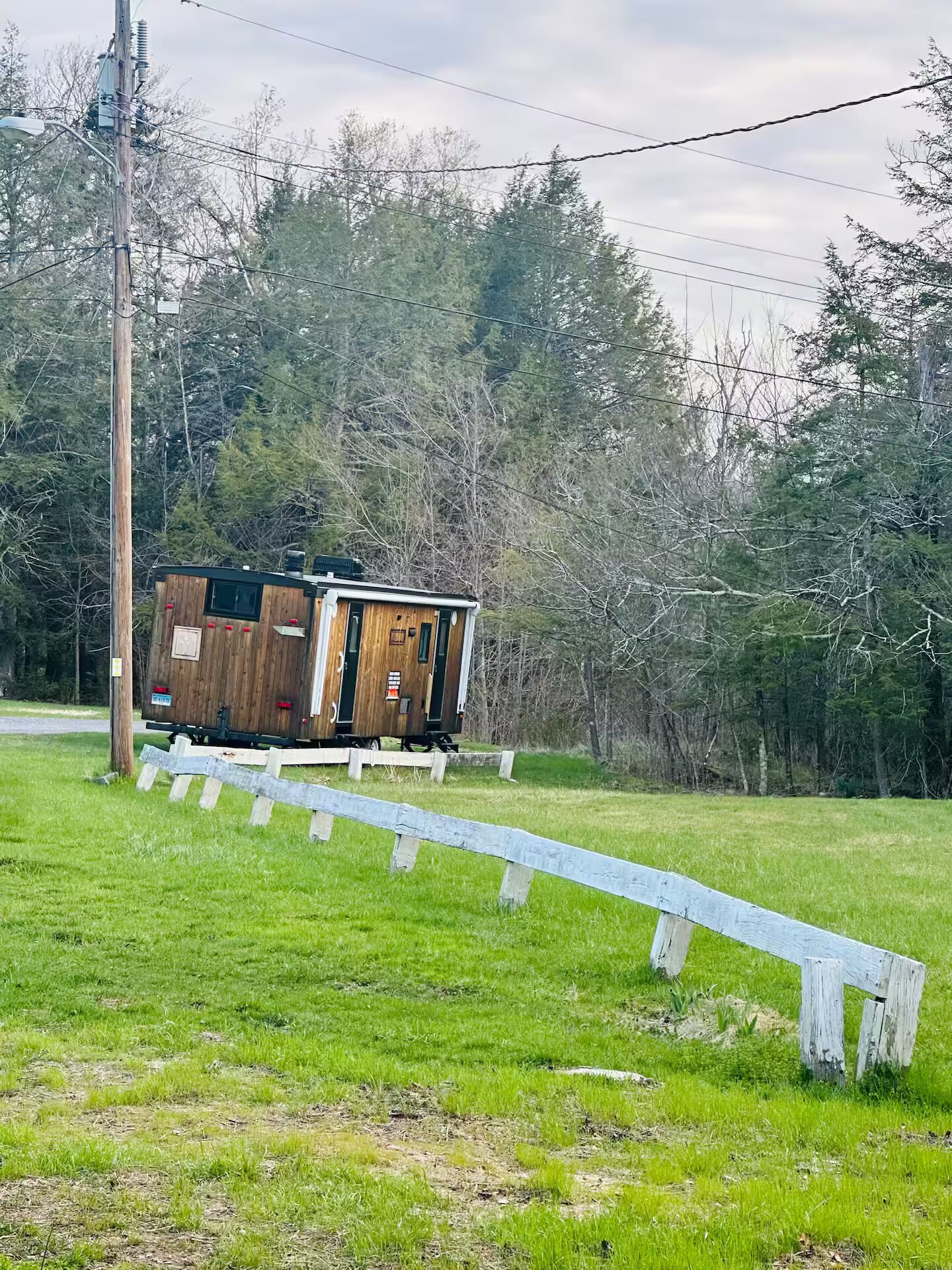 Een kleine houten hut met een metalen dak, gelegen op een grasveld met een houten omheining op de voorgrond.