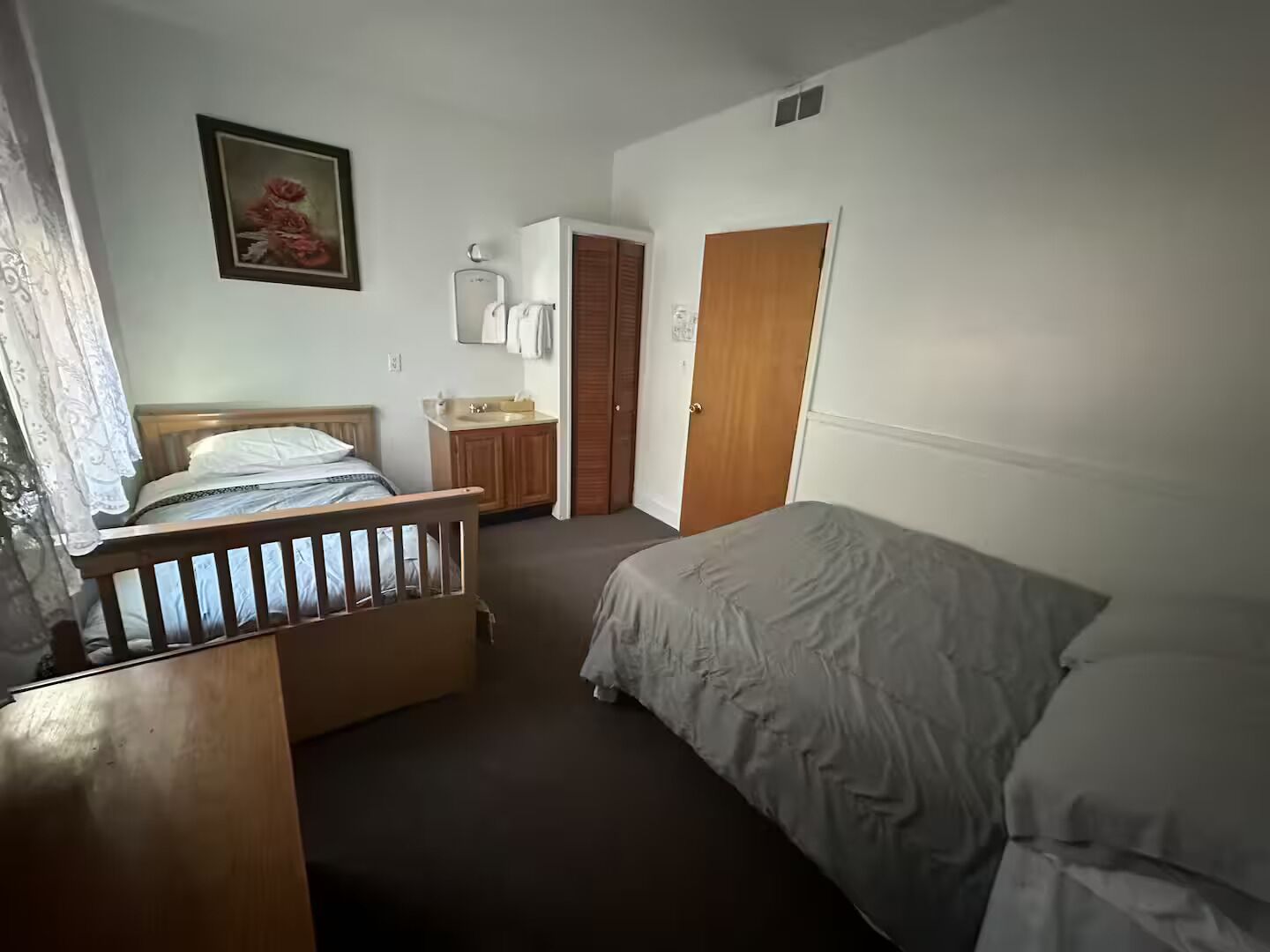 A bedroom with a wooden bed, a bedside table, a small dresser, a mirror, and a door leading to another room.