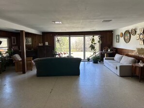 A spacious living room with a large green sofa, a white couch, and a wooden cabinet. There are potted plants and a large window providing a view of the outdoors.