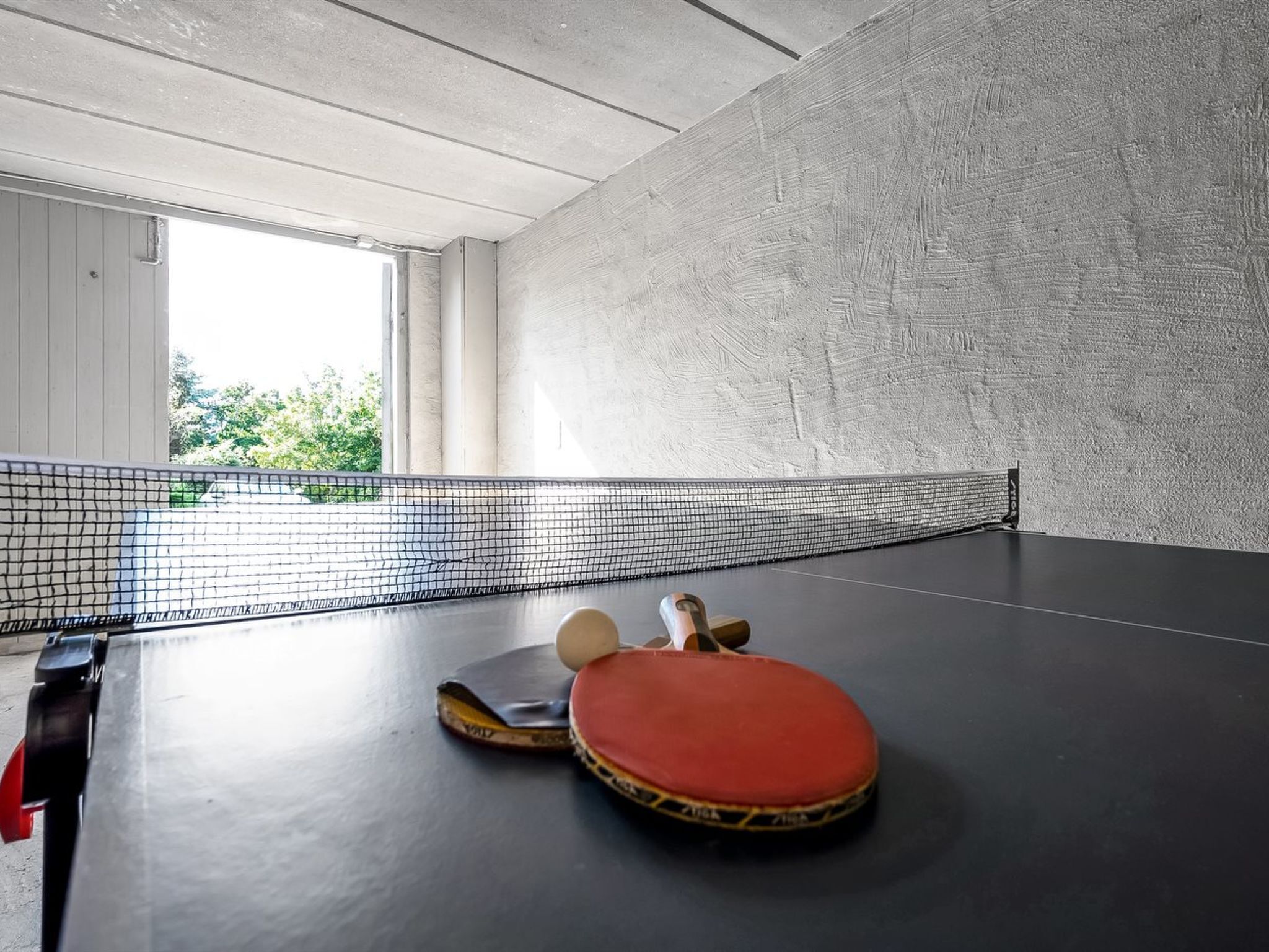 Une table de ping-pong avec des raquettes et une balle, une fenêtre donnant sur un paysage verdoyant et un mur à texture.