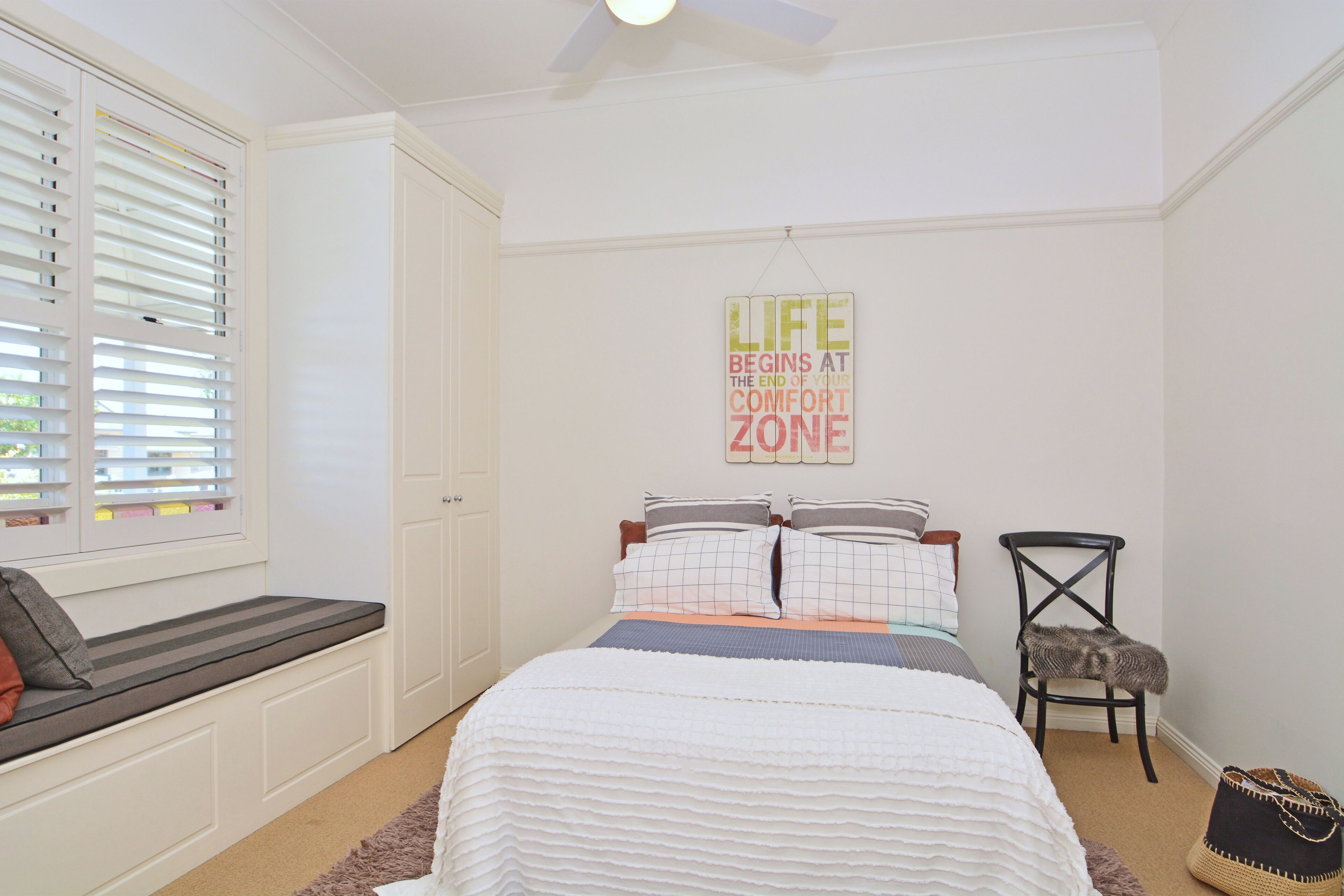 A bedroom with a bed, a bench, a chair, a wall sign, and a window with blinds.