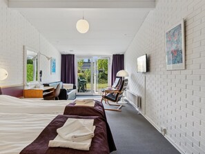 Ein Hotelzimmer mit zwei Betten, einem Schreibtisch, einem Sessel, einem Fernseher und einem Balkon mit Blick auf Grünflächen.