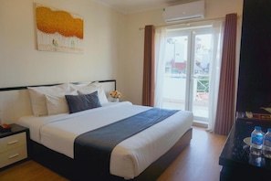 A hotel room with a bed, a nightstand, a desk with water bottles, an air conditioning unit, a window with curtains, and a painting on the wall. - Hanoi Serenity Hotel 2 (Hanoi)