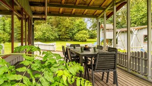 Un patio techado al aire libre con una mesa y sillas, rodeado por una terraza de madera y vistas a un parque infantil.