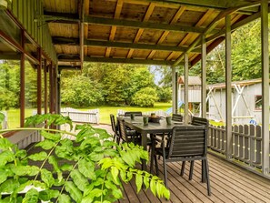Eine überdachte Terrasse im Freien mit Tisch und Stühlen, umgeben von einer Holzterrasse und Blick auf einen Spielplatz.