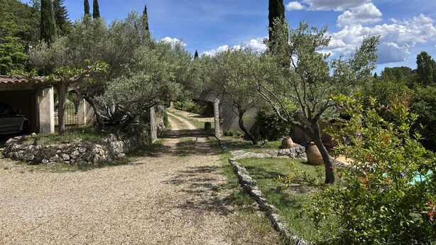 Un chemin de gravier traverse une oliveraie bordée de murs de pierre et mène à un bâtiment rustique.