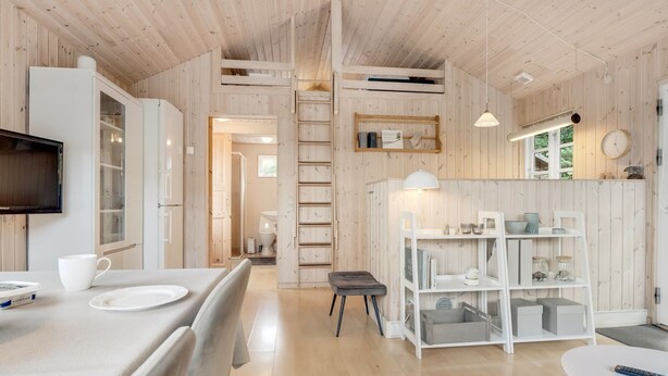 Een moderne, goed verlichte kamer met een houten plafond, een flatscreen televisie aan de muur, een eettafel met stoelen, een klein krukje en een witte kast met planken.