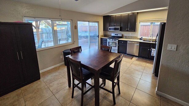 A kitchen with a dining table, chairs, a ceiling fan, and a view of the backyard through large windows. - Cozy house in the heart of Willow Springs (Rosamond)