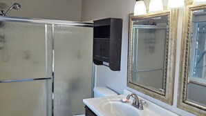 A bathroom with a toilet, two sinks, a shower, and a mirror with lights. - Cozy house in the heart of Willow Springs (Rosamond)